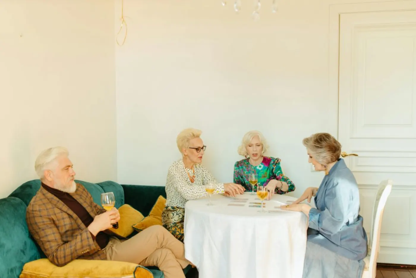 Elderly friends gather indoors, playing cards and enjoying wine in a cozy setting.