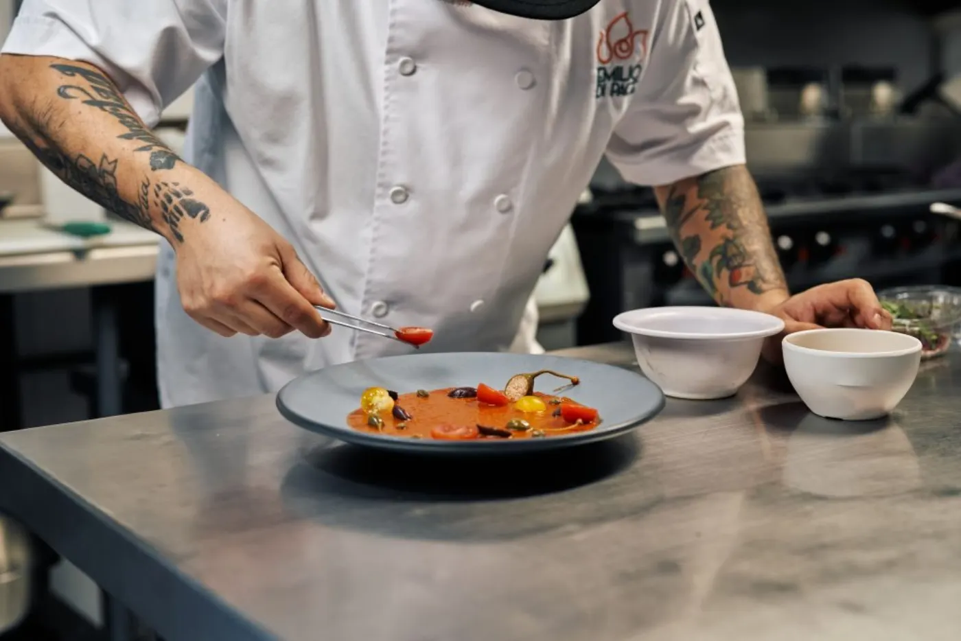 Tattooed chef garnishing a gourmet dish in a professional kitchen setting.