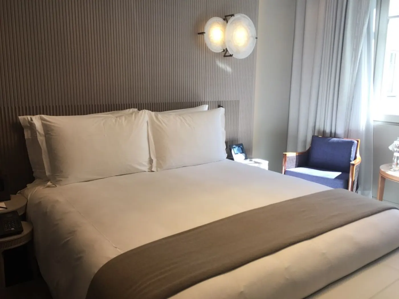Chambre du Lutetia avec lit blanc, plaid taupe, applique en forme de coquillage et fauteuil bleu près de la fenêtre