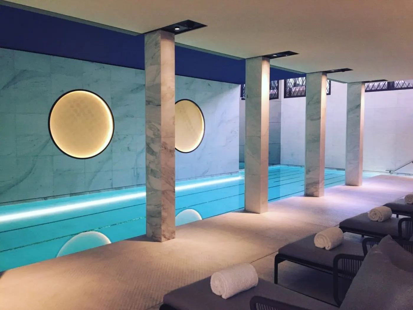 Piscine intérieure en marbre, colonnes, disques lumineux ronds et transats garnis de serviettes roulées