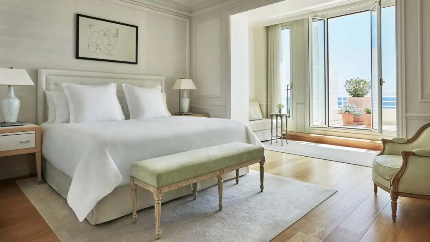 Chambre claire avec lit blanc, banquette vert amande et porte-fenêtre ouverte sur une terrasse face à la mer