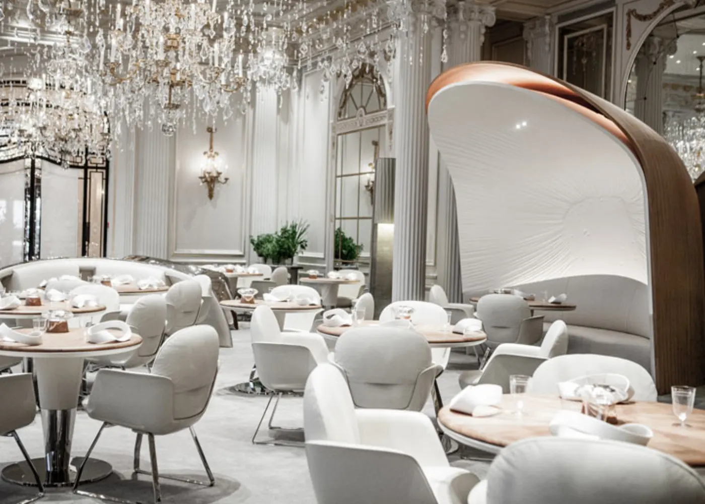 Salle de restaurant vide sous des lustres de cristal, fauteuils blancs et alcôve sculpturale en bois et toile