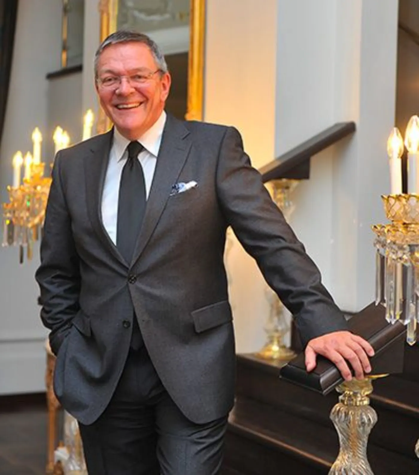 Ralph Radtke souriant en costume gris, appuyé à la rampe d'un escalier près de candélabres dorés