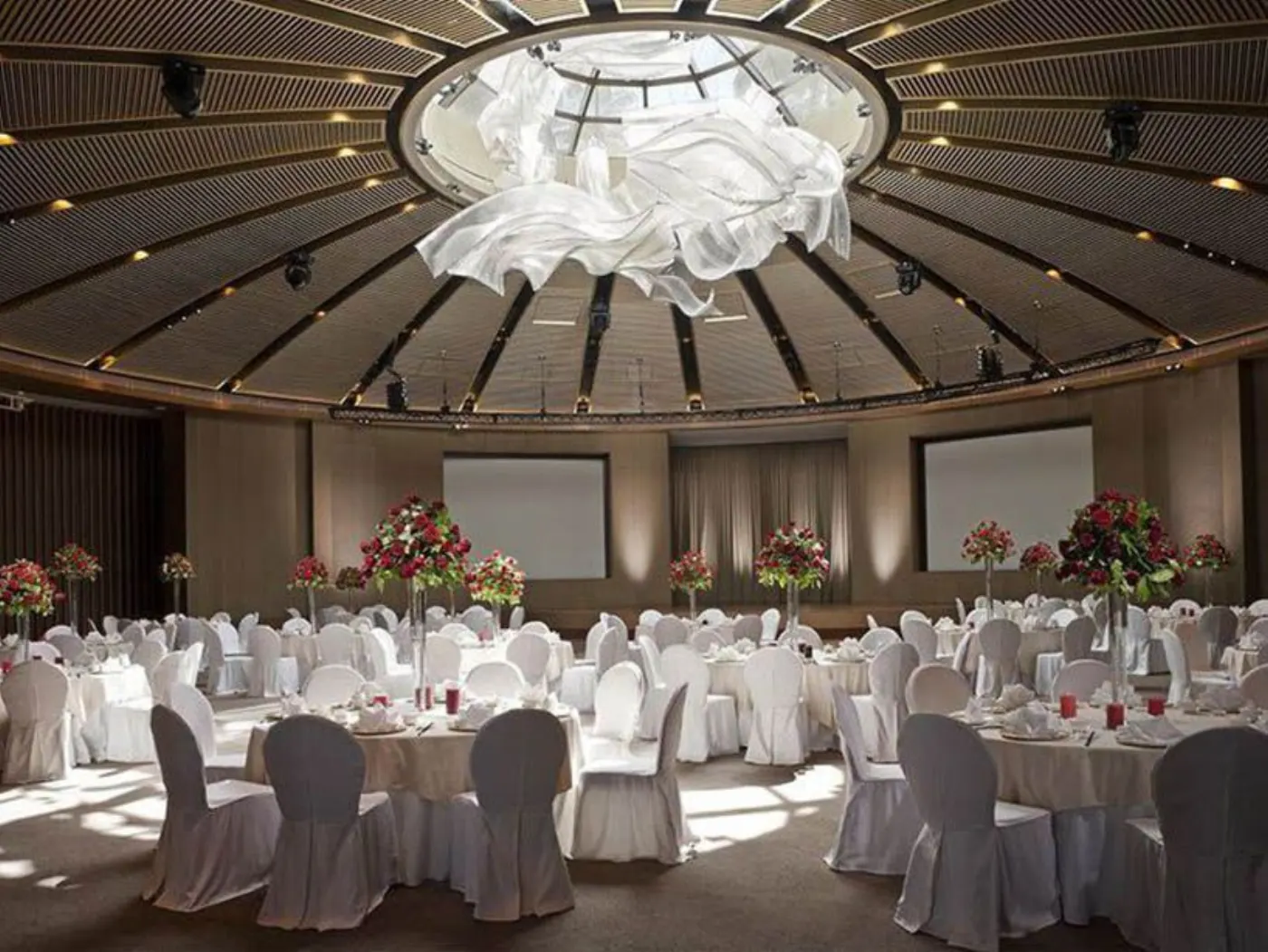 Salle de banquet circulaire sous un dôme à lattes, tables nappées de blanc et hautes compositions de roses rouges