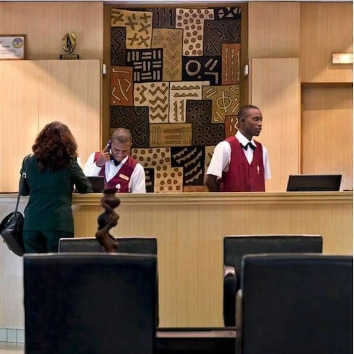 Comptoir de réception en bois clair, deux réceptionnistes en gilet rouge et une cliente devant une tenture africaine