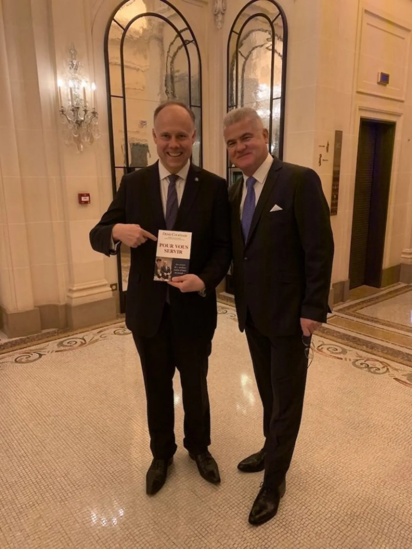 Deux hommes en costume debout dans le hall d'un palace, l'un tenant le livre Pour vous servir de Denis Courtiade