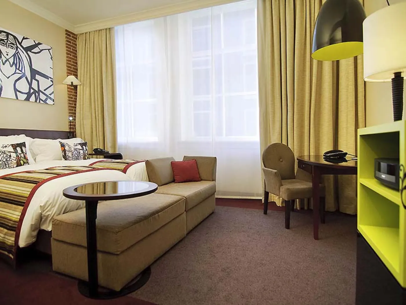 Chambre d'hôtel avec lit, banquette beige, rideaux jaunes et meuble vert anis devant une fenêtre voilée