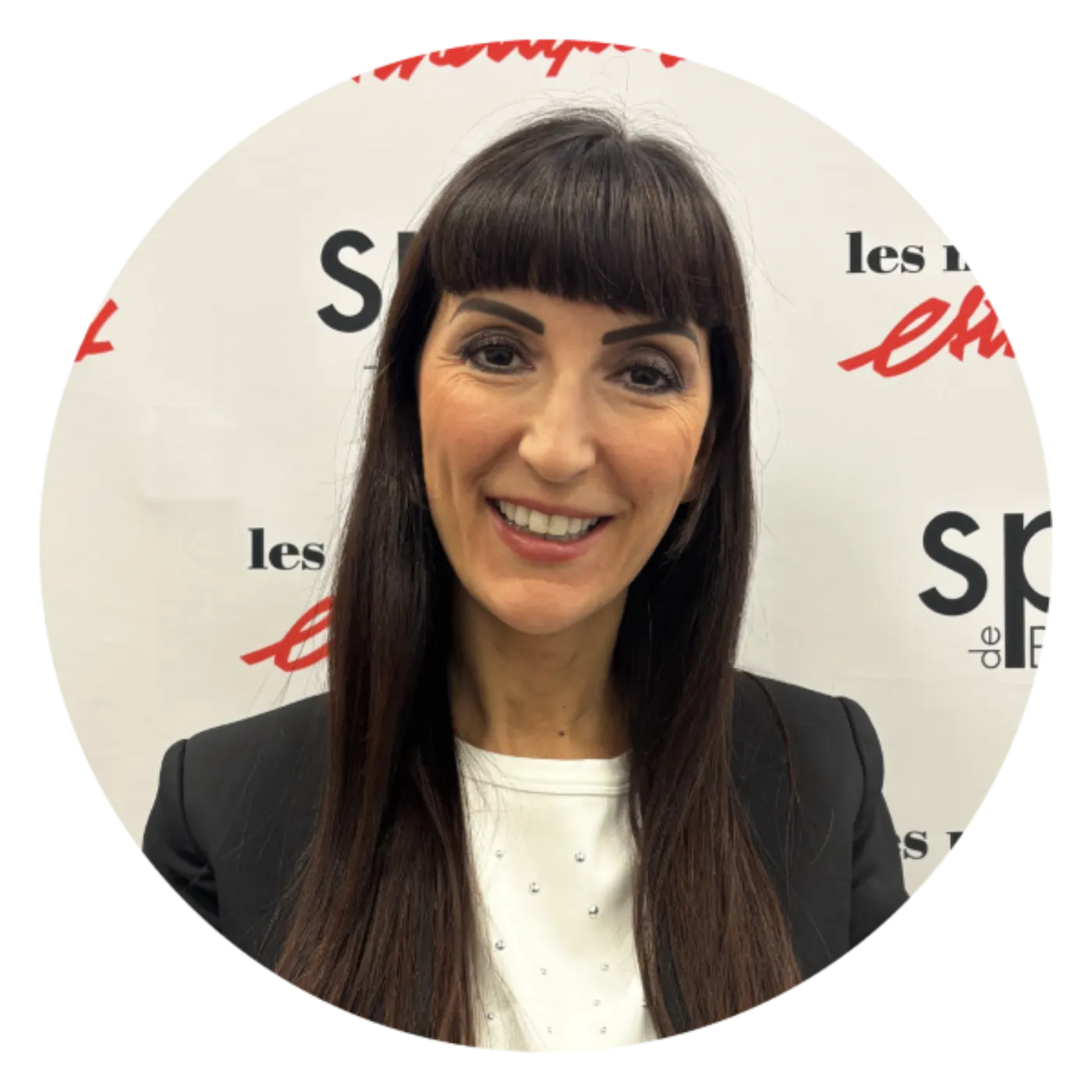 Femme brune souriante en blazer noir, devant un mur de presse Les Nouvelles Esthétiques Spa