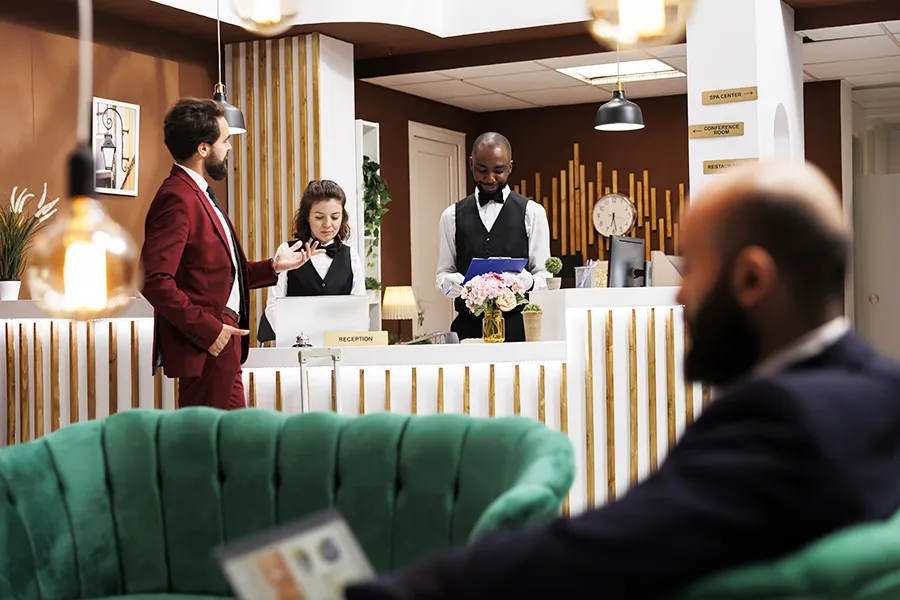 Scène de réception d'hôtel : un client accueilli au comptoir pendant qu'un homme observe depuis un canapé