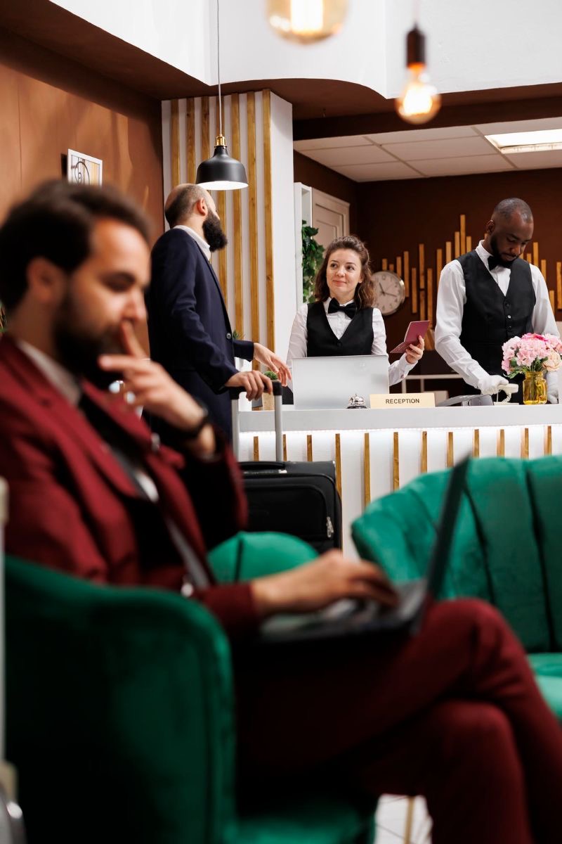 Hotel reception at work: front-desk staff greeting an arriving guest while another guest works from a sofa