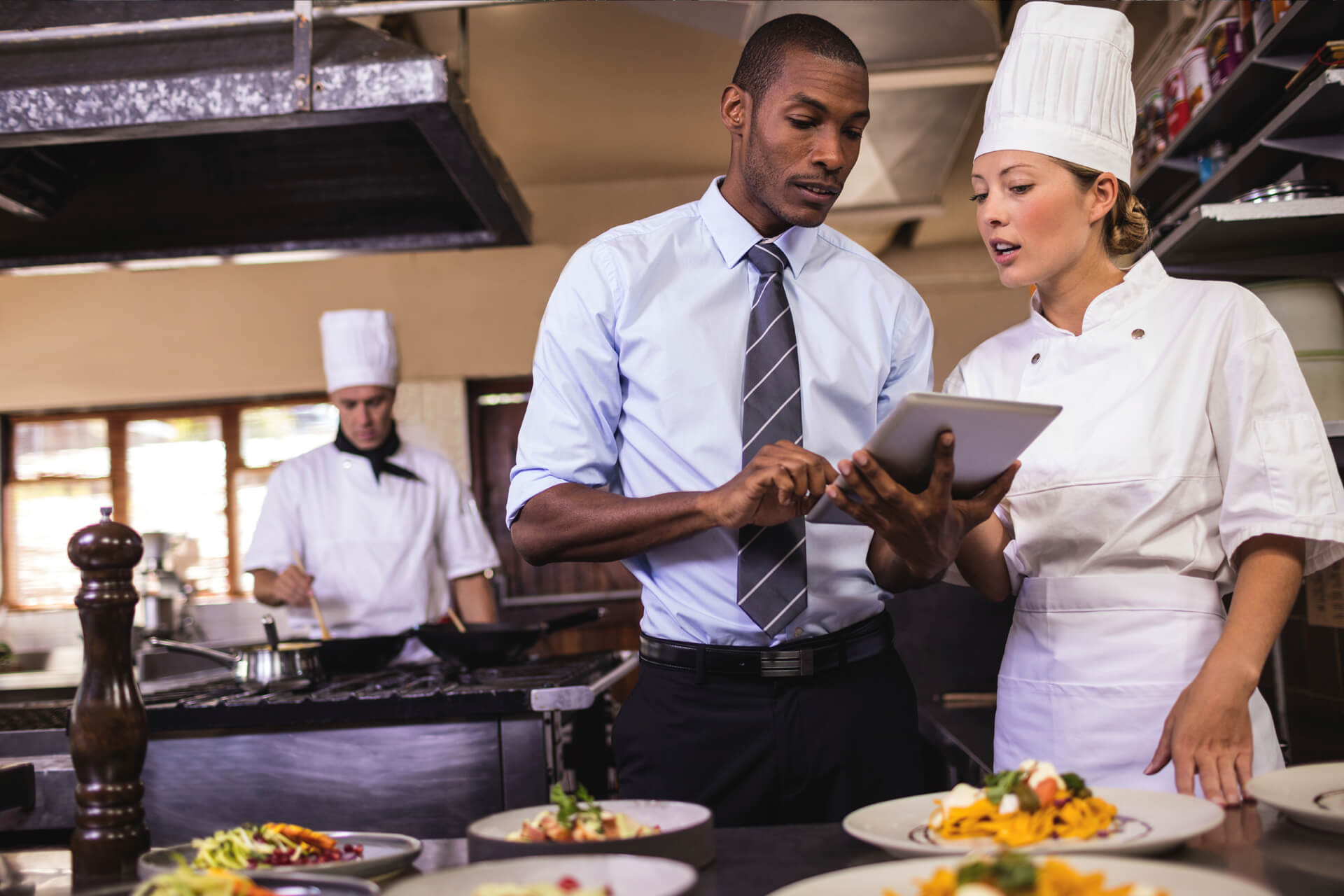 Cuisine professionnelle : un responsable montre une tablette à une cheffe en toque, assiettes dressées au premier plan