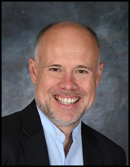 Portrait of Laurent Delporte in a black jacket and light blue shirt against a blue backdrop