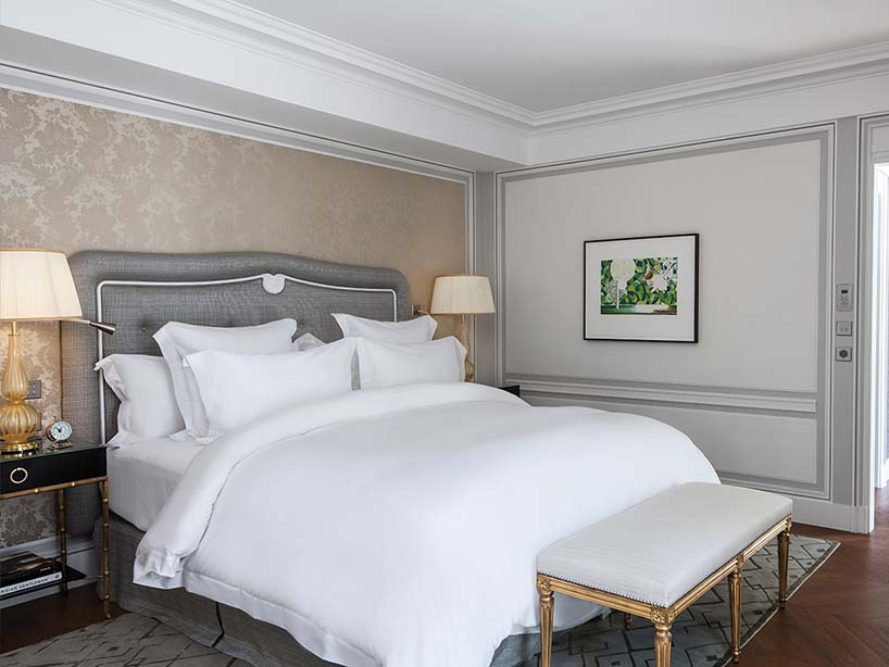 Chambre de l'Hôtel de Crillon : tête de lit grise capitonnée, literie blanche et banquette dorée au pied du lit