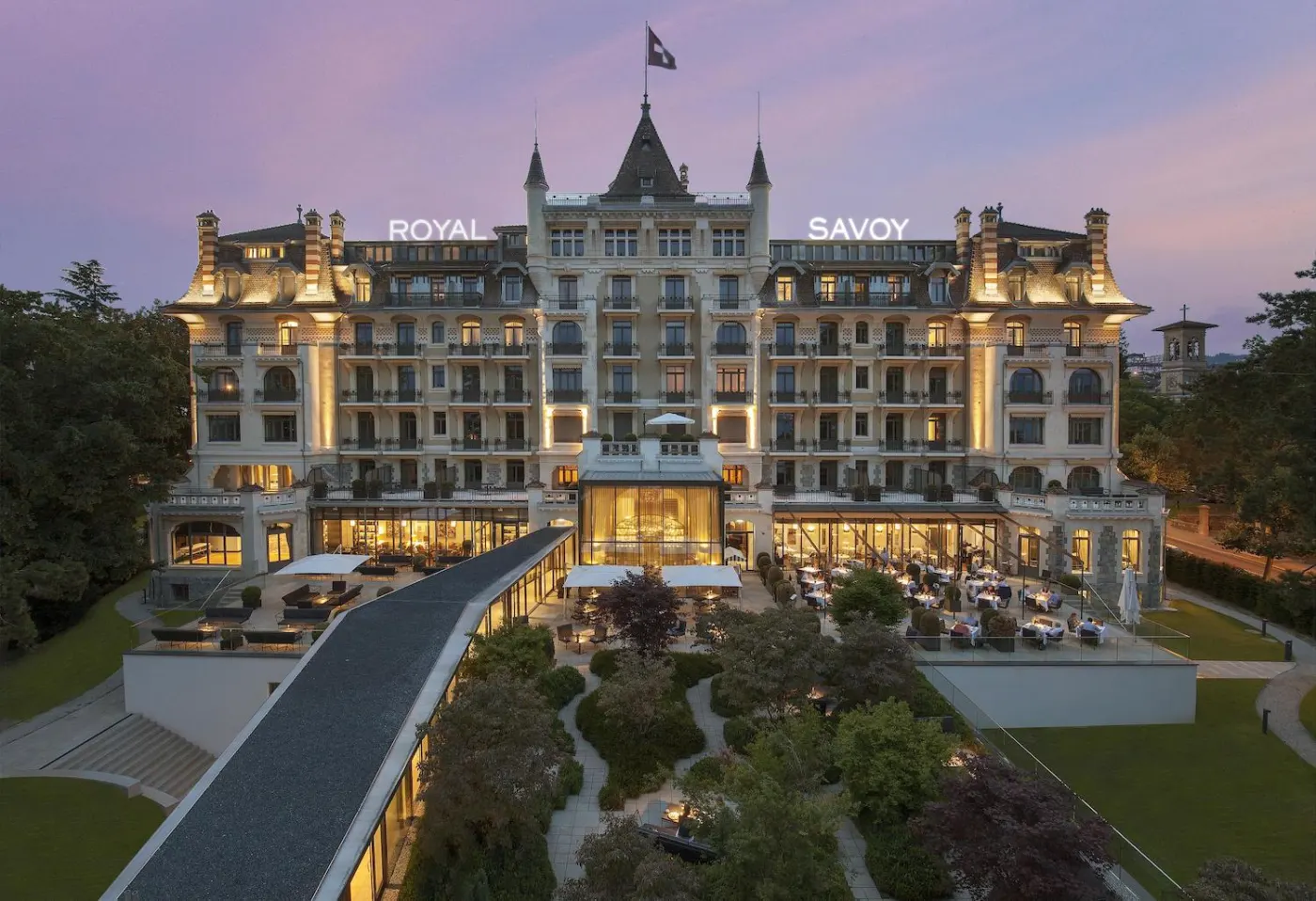 The Royal Savoy lit up at dusk, rooftop signage, Swiss flag and garden-side restaurant terrace