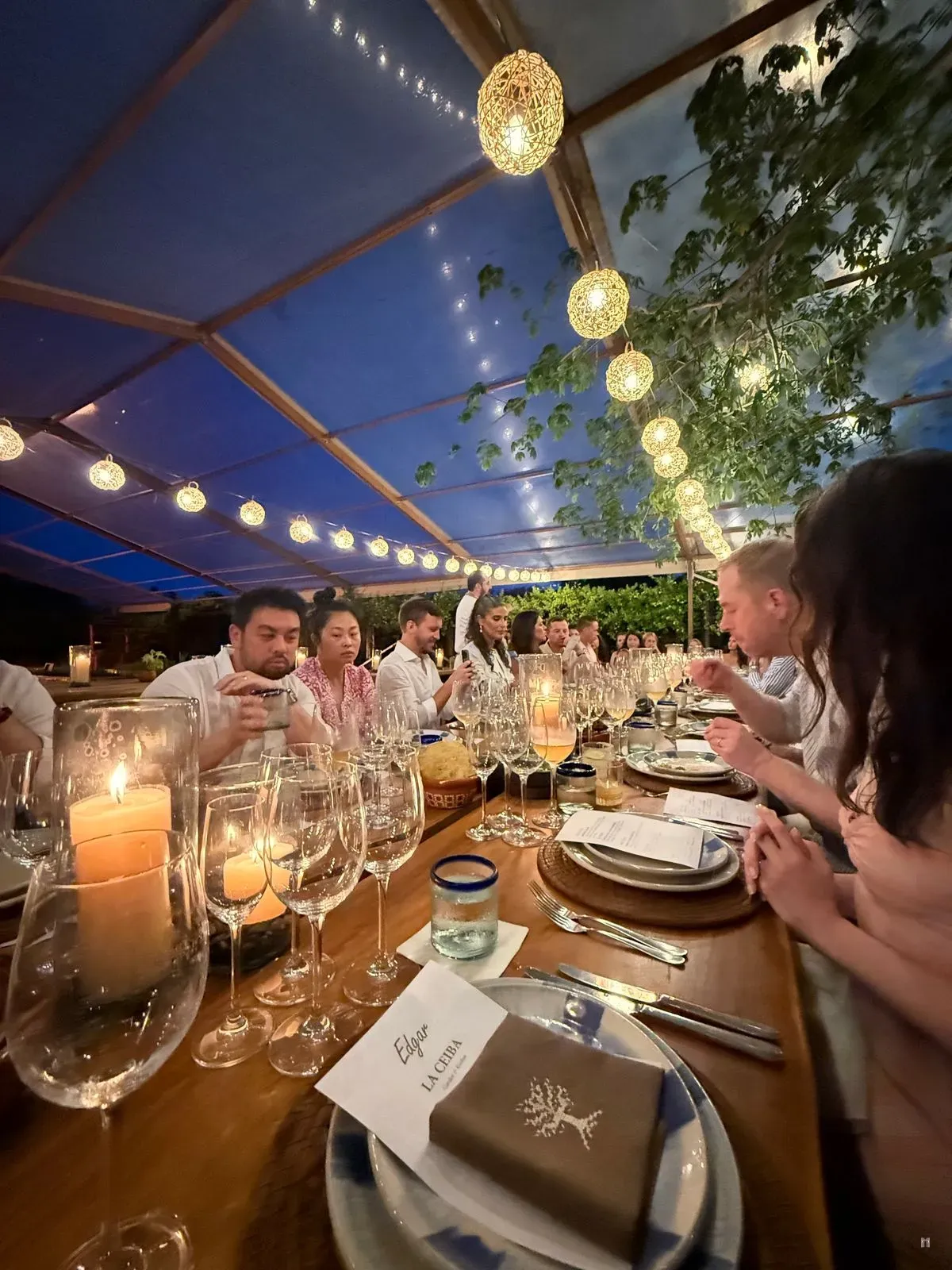 Grande table dressée sous une verrière et des lanternes lumineuses, avec bougies, verres et convives attablés, au dîner LA CEIBA GARDEN & KITCHEN
