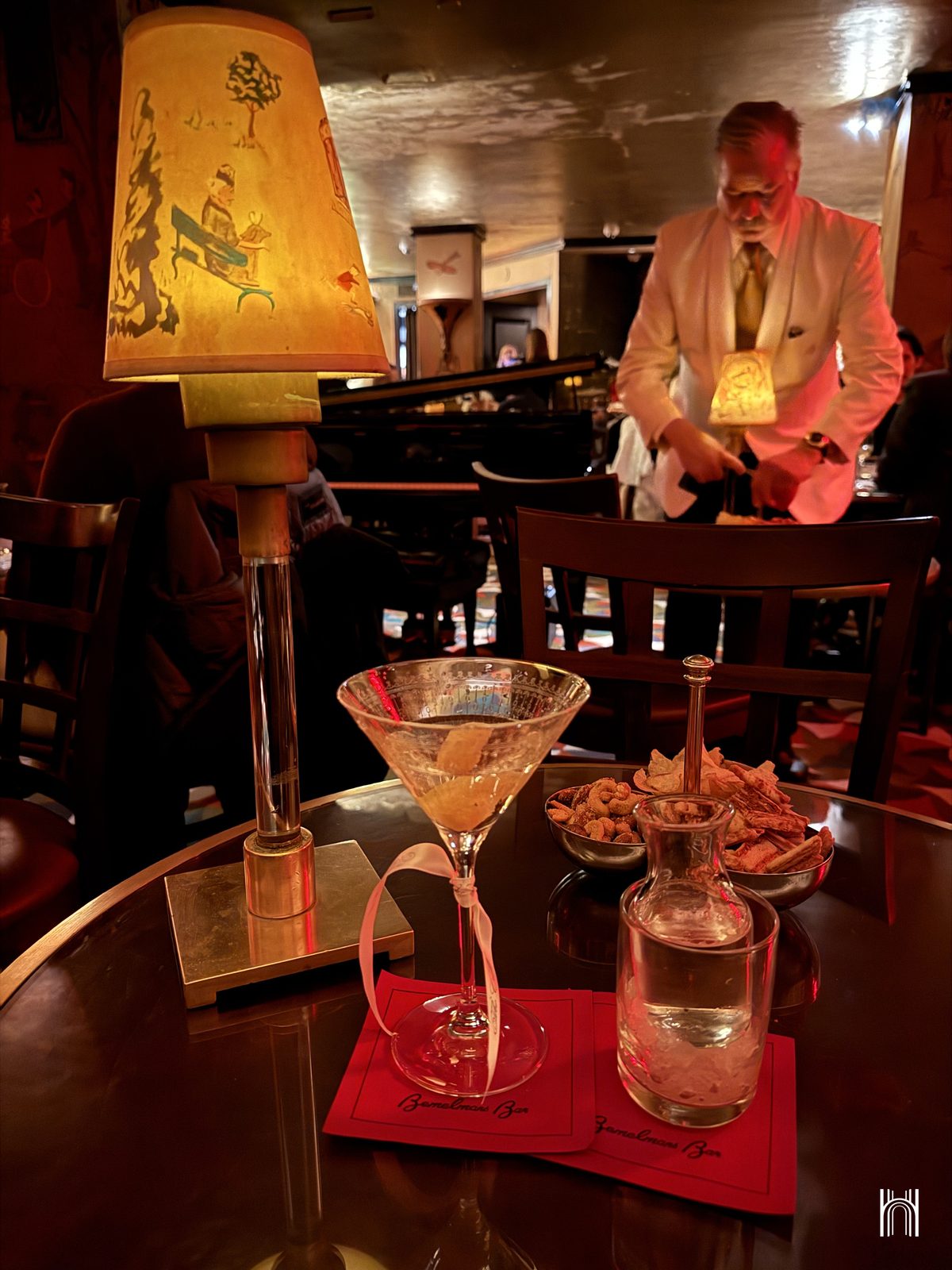 Un martini et une lampe aux abat-jour dessinés au Bemelmans Bar, avec un serveur en veste blanche en arrière-plan