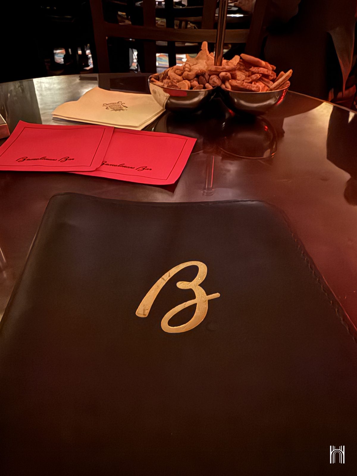 Table du Bemelmans Bar avec sous-verres rouges, serviettes et coupelles de grignotage, devant une carte au monogramme B doré