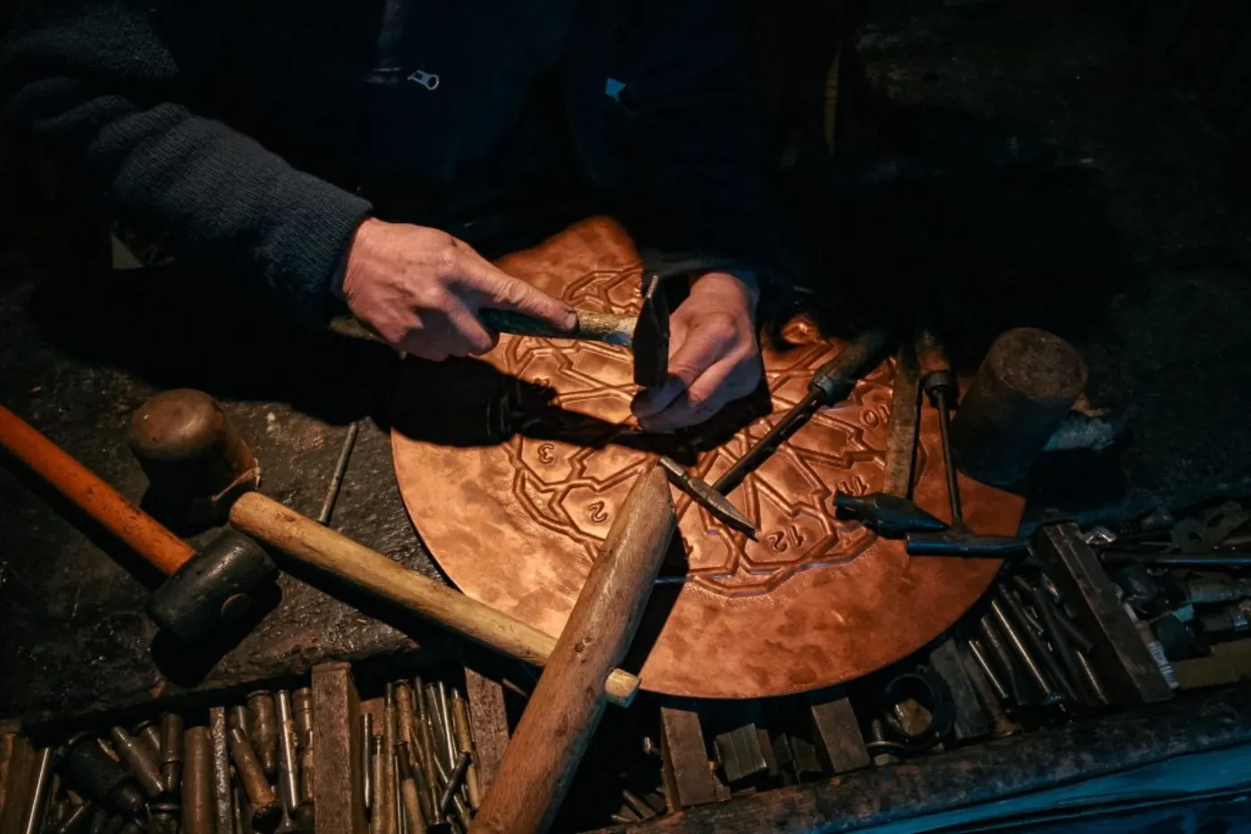 An artisan meticulously crafting a detailed metal plate using traditional tools in a dimly lit workshop.