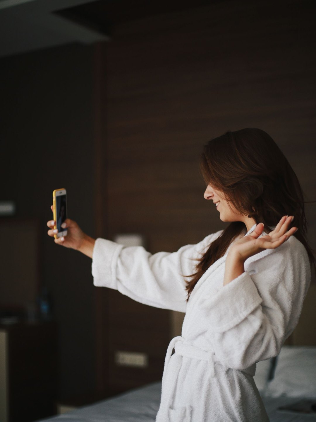 Young woman in a white bathrobe taking a picture of herself with her smartphone in a hotel room, a symbol of a generation that shares its places on social media